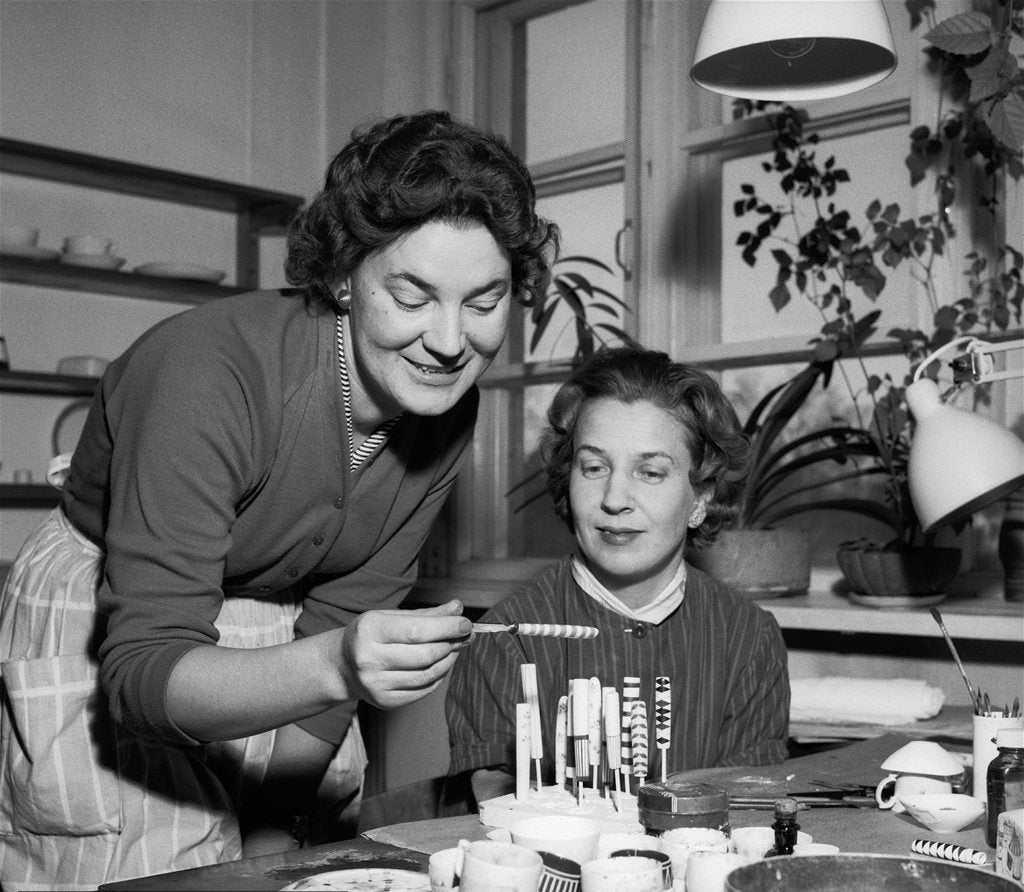 装飾した細い試作片を手にする女性と見つめる女性の白黒写真｜机に絵の具と器が並ぶ